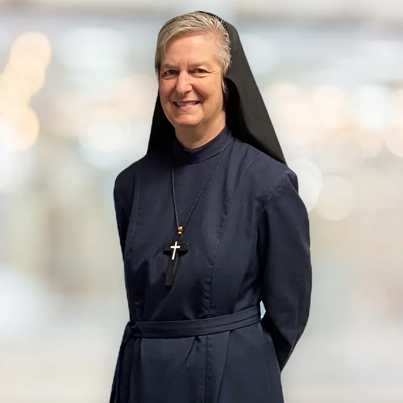 A smiling nun in a traditional black habit and veil, wearing a crucifix necklace, standing in front of a softly blurred background.