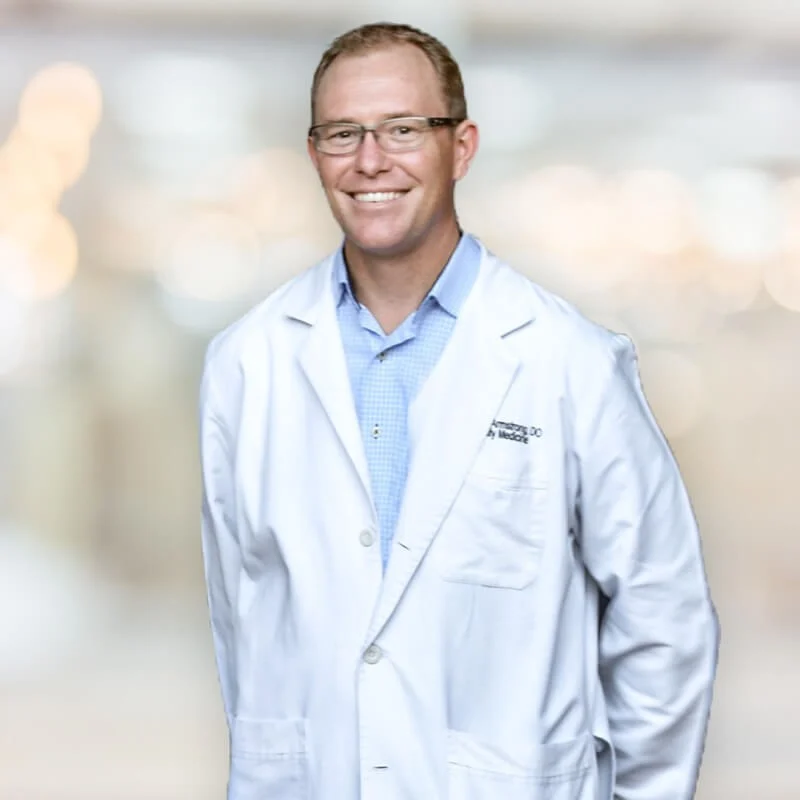 A smiling middle-aged man in glasses wearing a white lab coat with an embroidered name and title, standing in front of a softly blurred background.