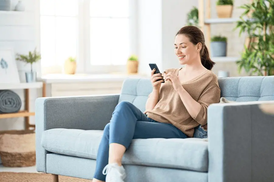Woman smiling while using smartphone, sitting comfortably on a blue sofa in a bright living room with plants.