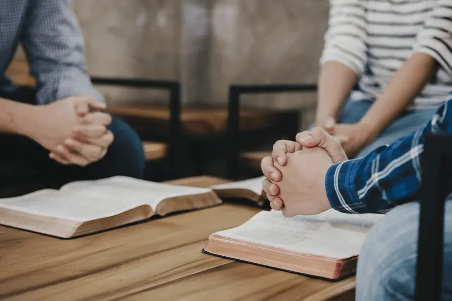 Two people holding hands over a table with open bibles during a group study session.