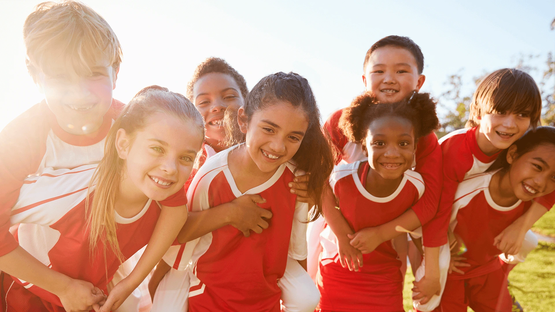 A group of diverse children in red sports uniforms smiling and embracing each other in a sunny outdoor setting.
