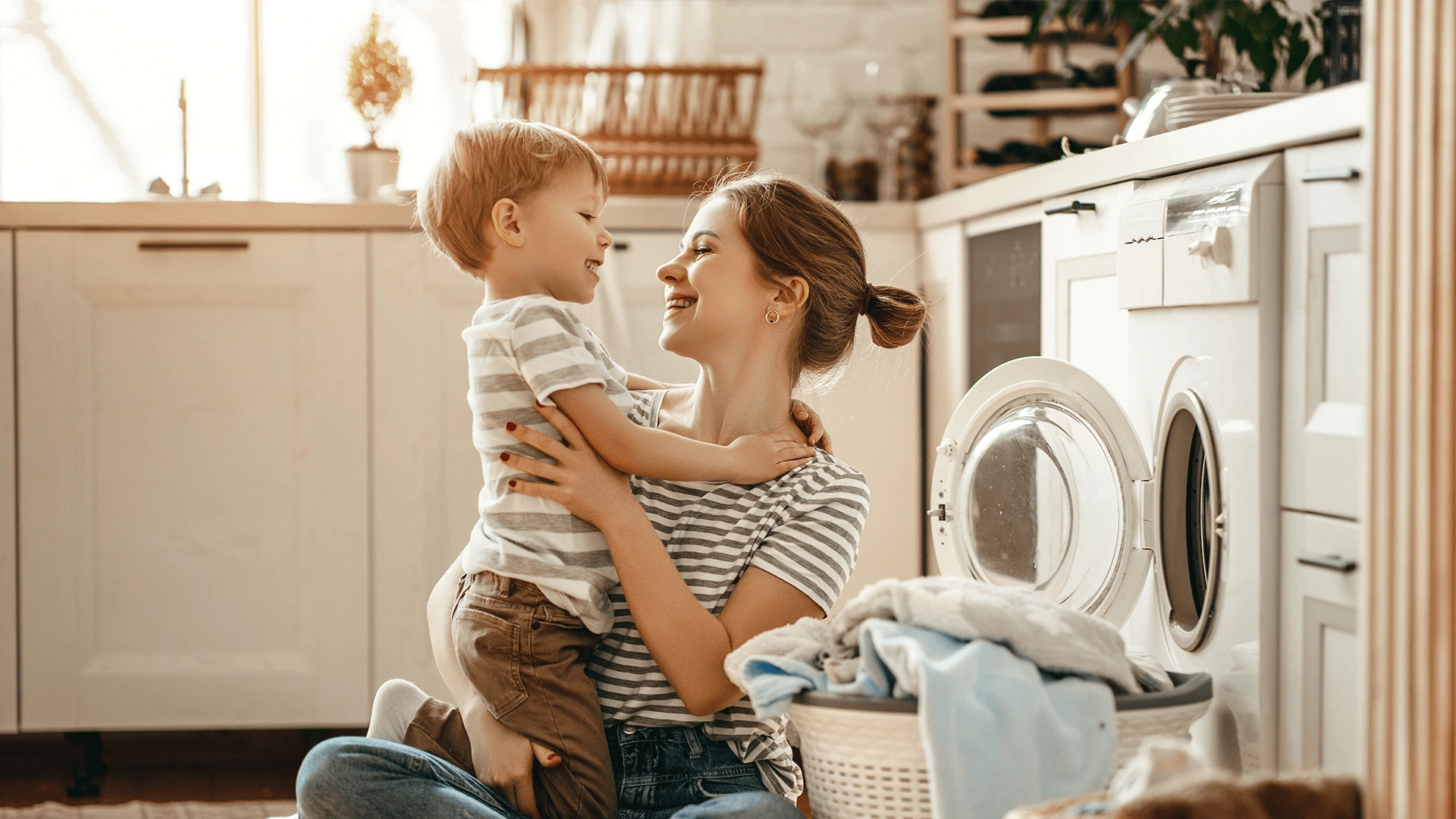A mother holding her toddler in a laundry room, both smiling at each other, with a washing machine and laundry basket nearby.