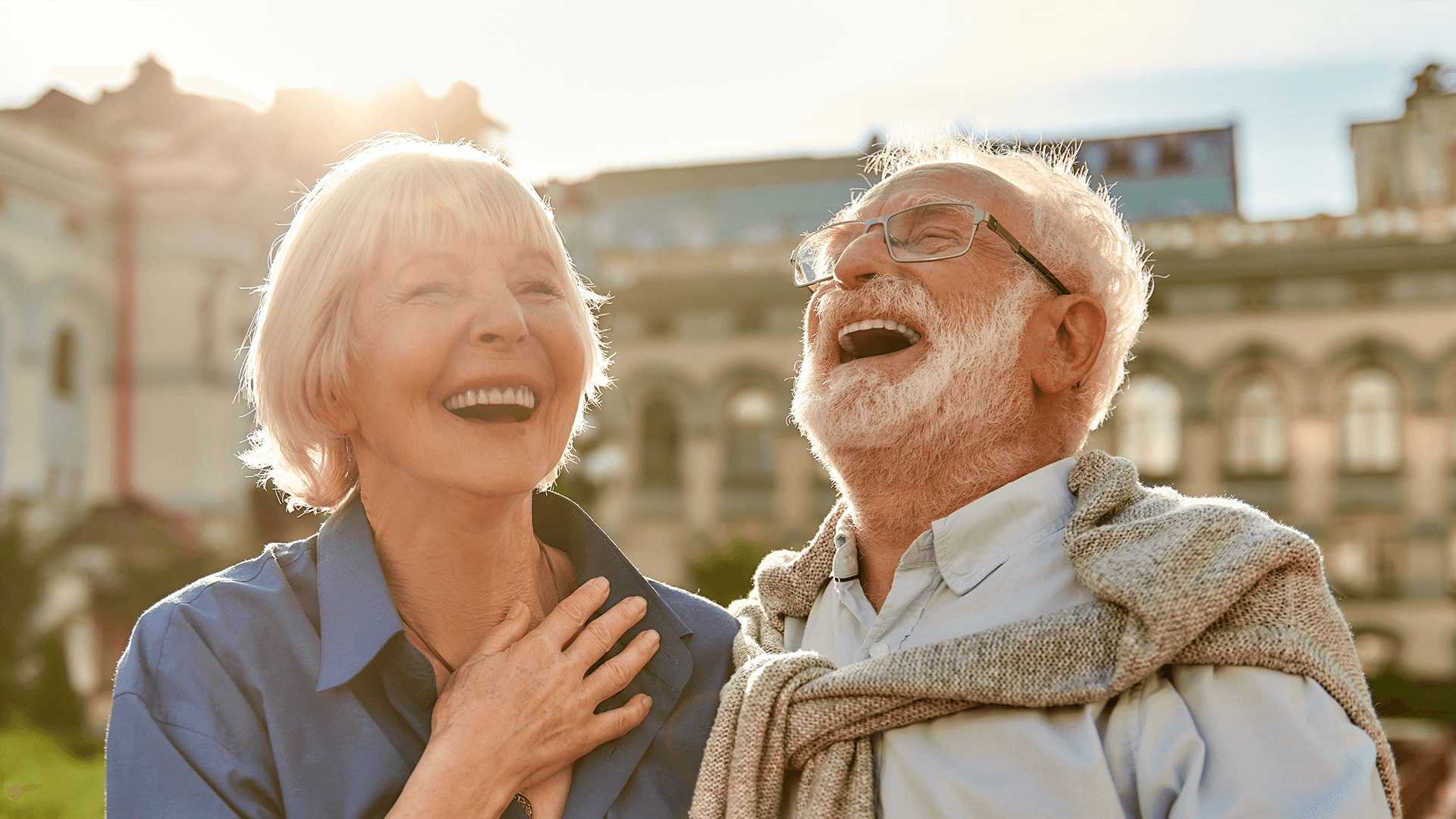 Elderly couple laughing joyfully together in a sunlit urban setting, with historical buildings in the background.