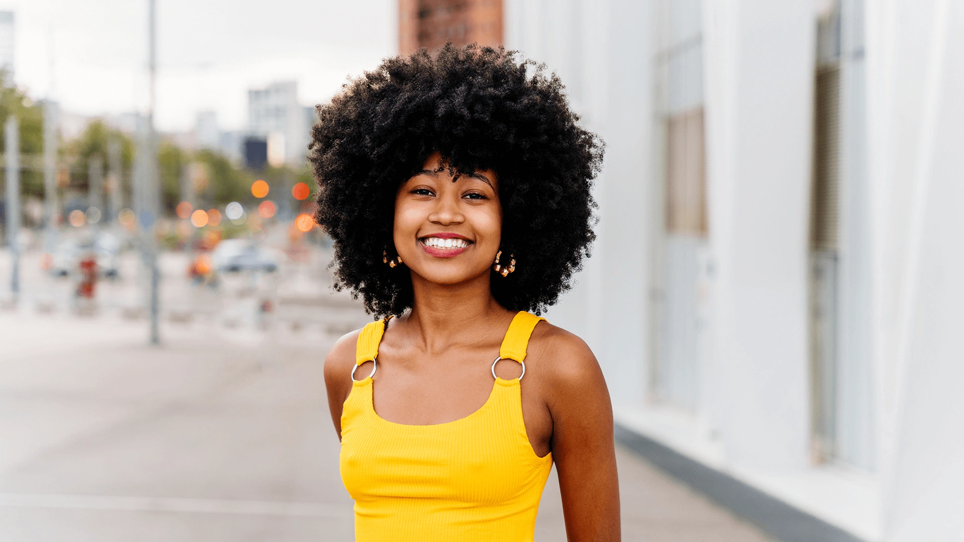 A joyful young woman with curly hair wearing a yellow tank top smiles brightly on a city street.