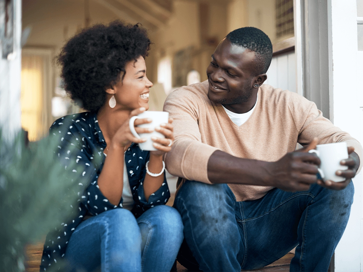 A young couple sits on a porch, smiling and holding mugs, deeply engaged in a joyful conversation.
