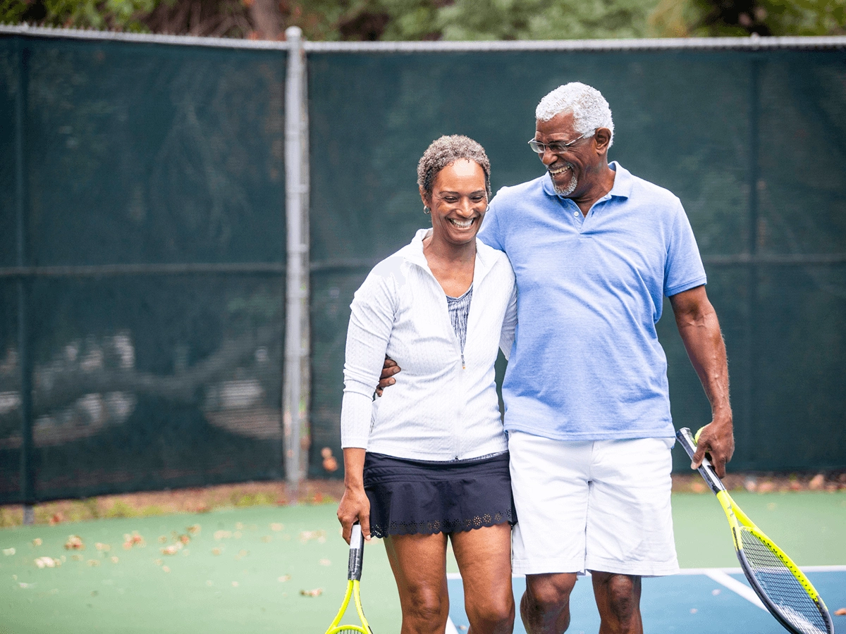 An older african american couple smiling and walking arm-in-arm on a tennis court, holding tennis rackets.