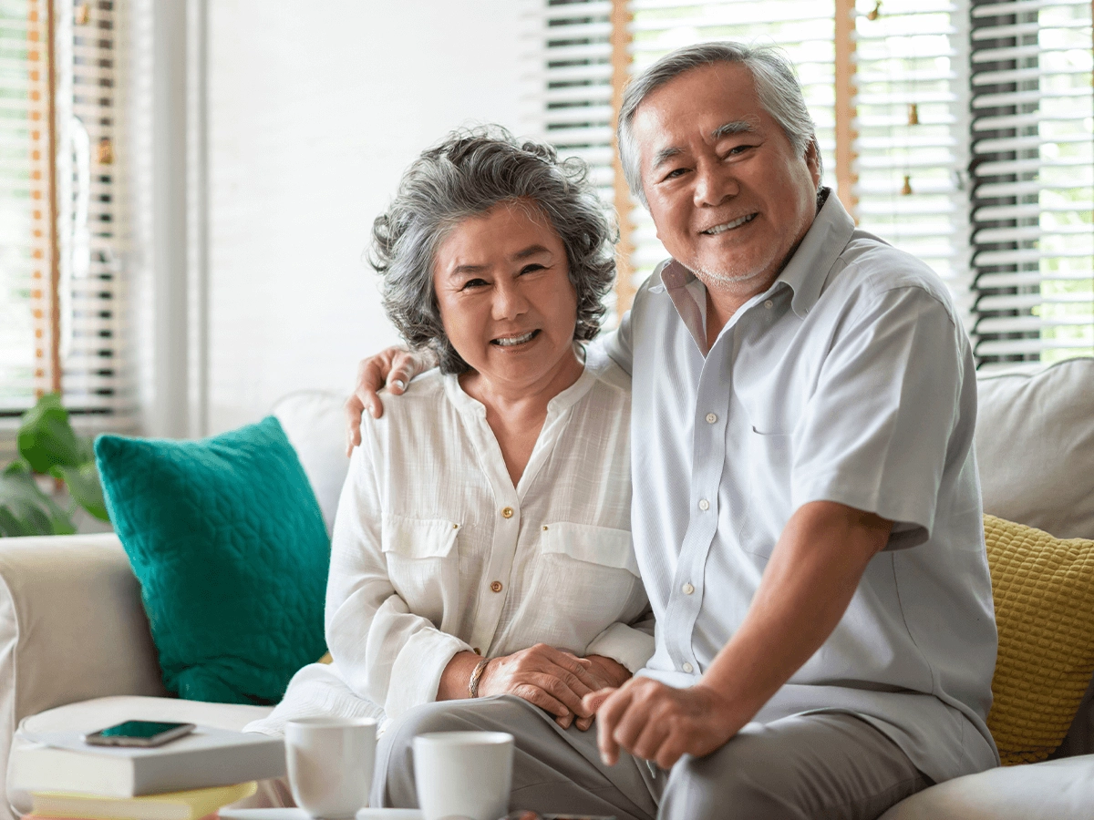 Elderly couple smiling and sitting closely together on a couch in a bright, plant-filled living room.