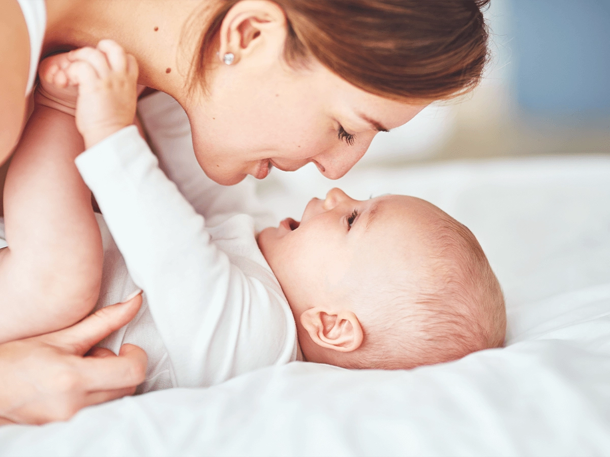 A mother affectionately looking at her baby who is lying on a bed, both engaging in a tender moment.