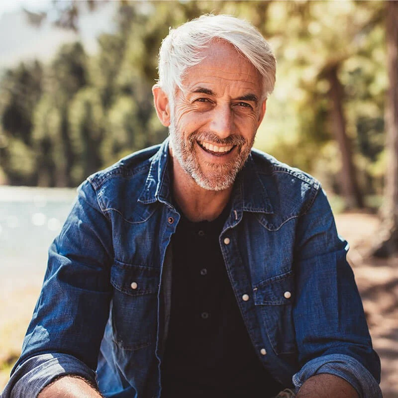 A smiling elderly man with white hair and a beard, wearing a denim shirt, sitting outdoors with trees and a lake in the background.