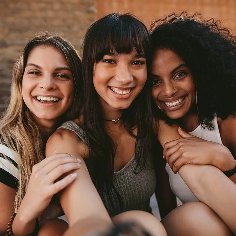 Three joyful young women of diverse ethnicities taking a close-up selfie, smiling broadly at the camera.