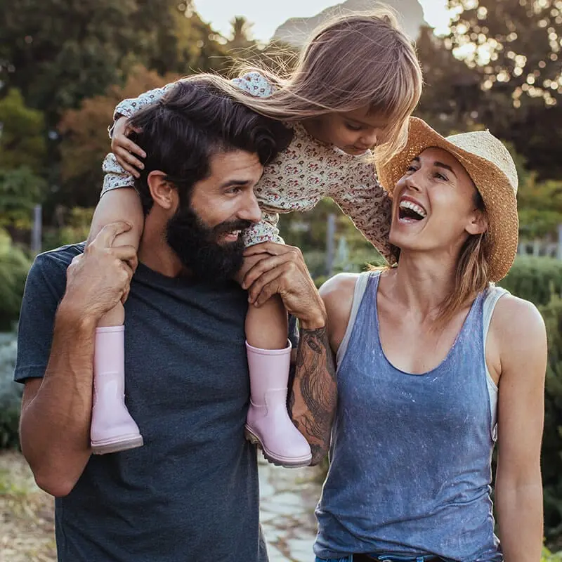 A happy family outdoors: a bearded man with a young girl on his shoulders and a laughing woman beside them, all in casual summer clothing.